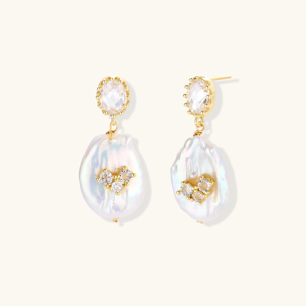 Gold-plated drop earrings with iridescent baroque pearls and crystal flower accents on cream background
