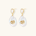 Gold-plated drop earrings with iridescent baroque pearls and crystal flower accents on cream background
