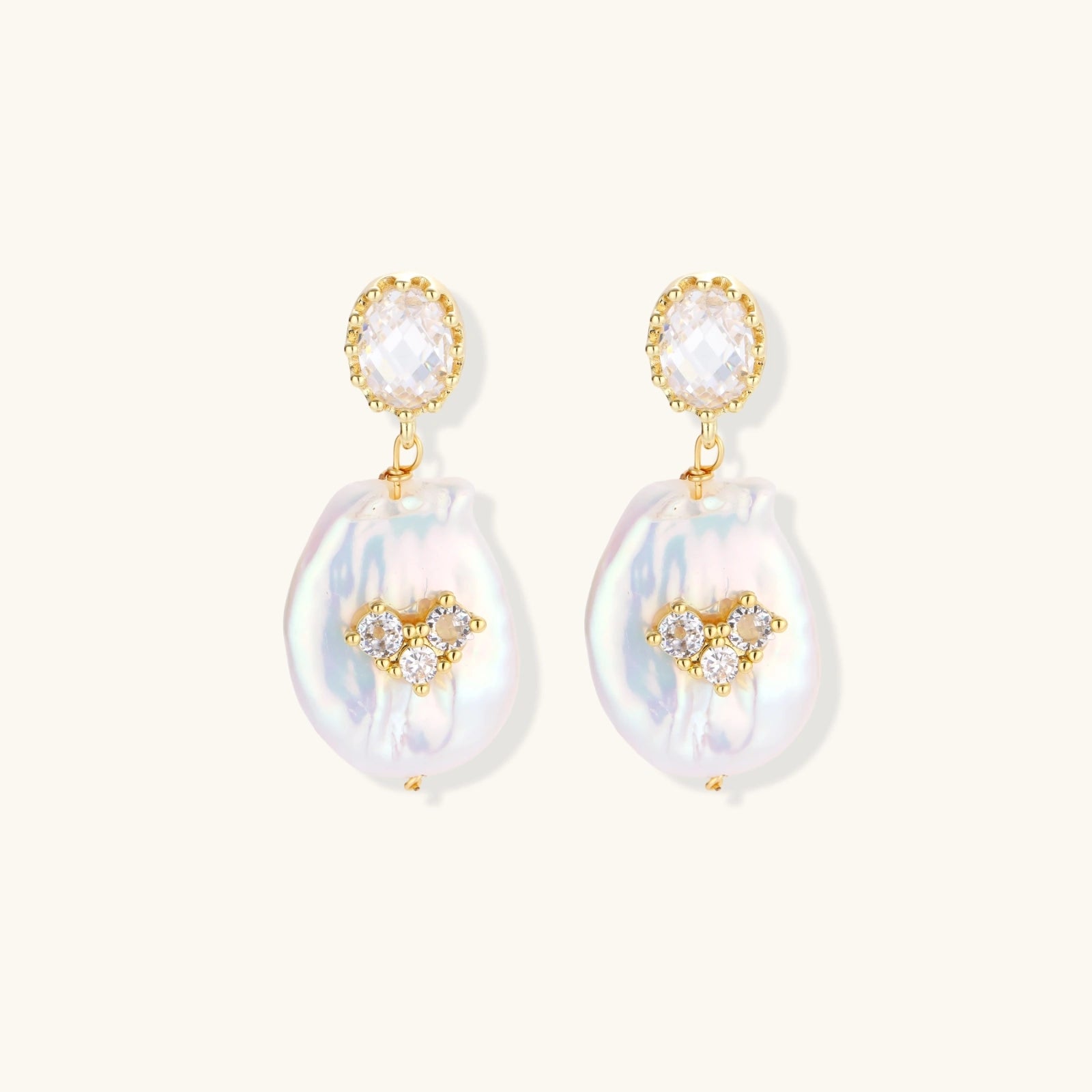 Gold-plated drop earrings with iridescent baroque pearls and crystal flower accents on cream background