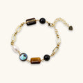 Gold chain bracelet with tiger's eye, obsidian, pearl, and iridescent abalone stones on cream background.