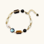 Gold chain bracelet with tiger's eye, obsidian, pearl, and iridescent abalone stones on cream background.