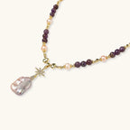 Gold necklace with iridescent baroque pearl pendant, purple amethyst beads, and crystal star charm on cream background