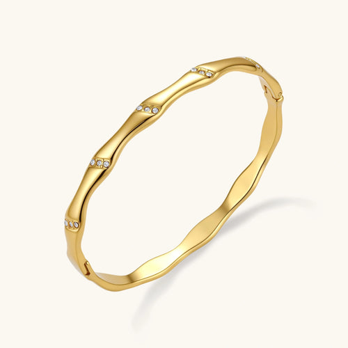 Gold-toned wavy bangle with bamboo-style segments and CZ crystal clusters on cream background. 18K plated stainless steel.