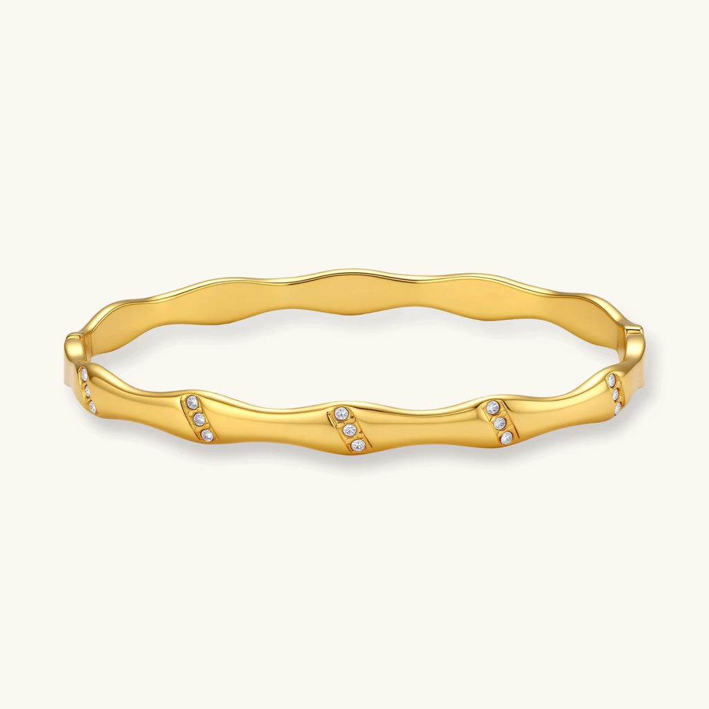 Gold-toned wavy bangle with bamboo-style segments and CZ crystal clusters on cream background. 18K plated stainless steel.