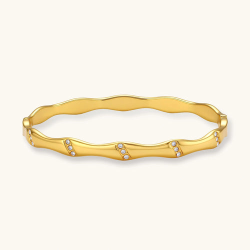 Gold-toned wavy bangle with bamboo-style segments and CZ crystal clusters on cream background. 18K plated stainless steel.