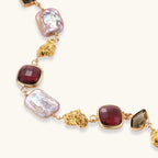 18k gold-plated copper bracelet with burgundy stones, smoky quartz, baroque pearls and ornate golden charms on white background.