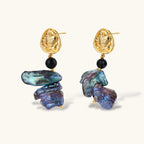 Ornate gold-plated drop earrings with black beads and iridescent blue-purple baroque pearls against white background.
